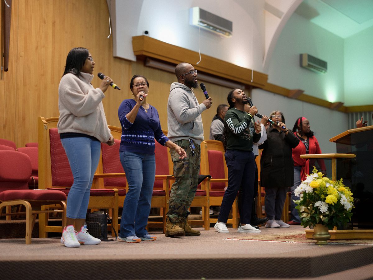 A row of people stand and sing at the front of a red-carpeted church sanctuary, some with microphones.