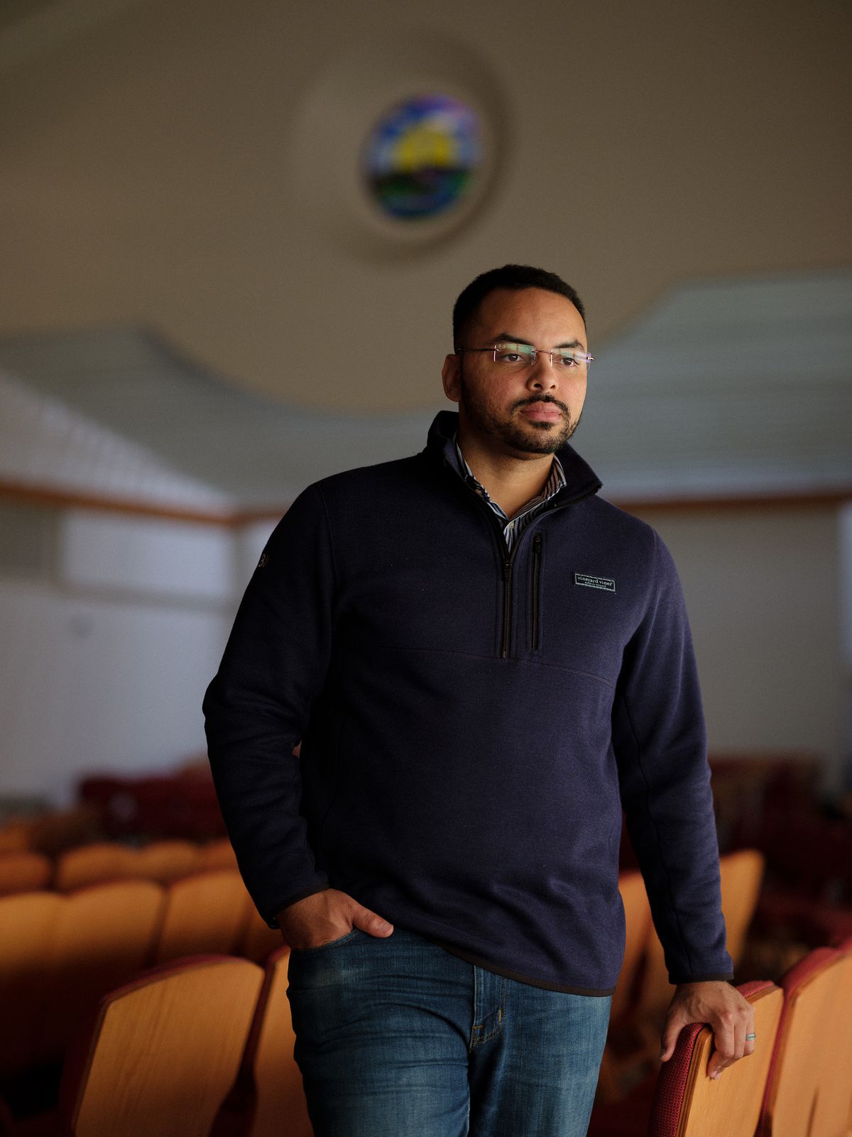 A younger Black man with glasses and wearing a blue sweater and jeans, standing among chairs in a church sanctuary.