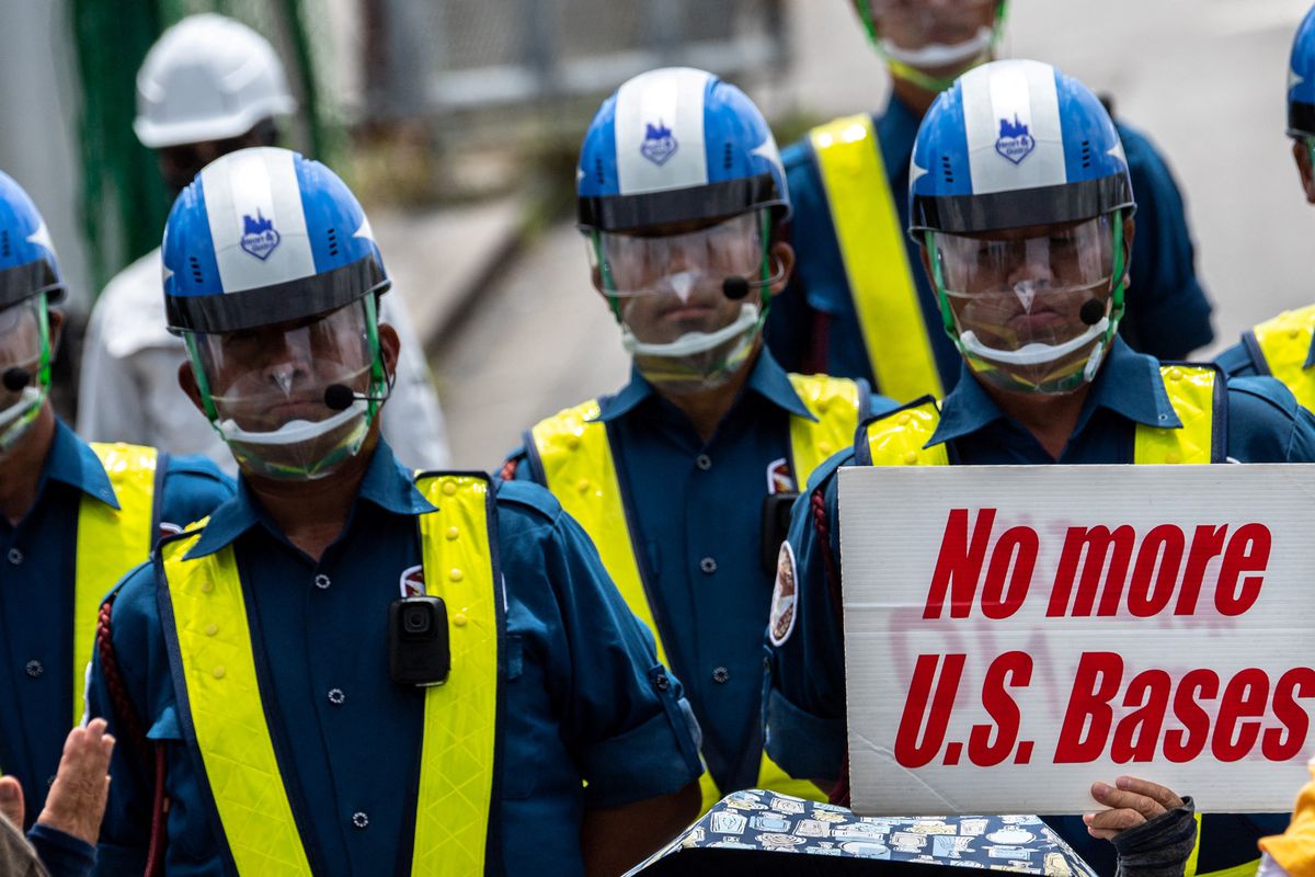 Uniformed men with helmets stand in a line, while a “No more US bases” sign can is held in the foreground.