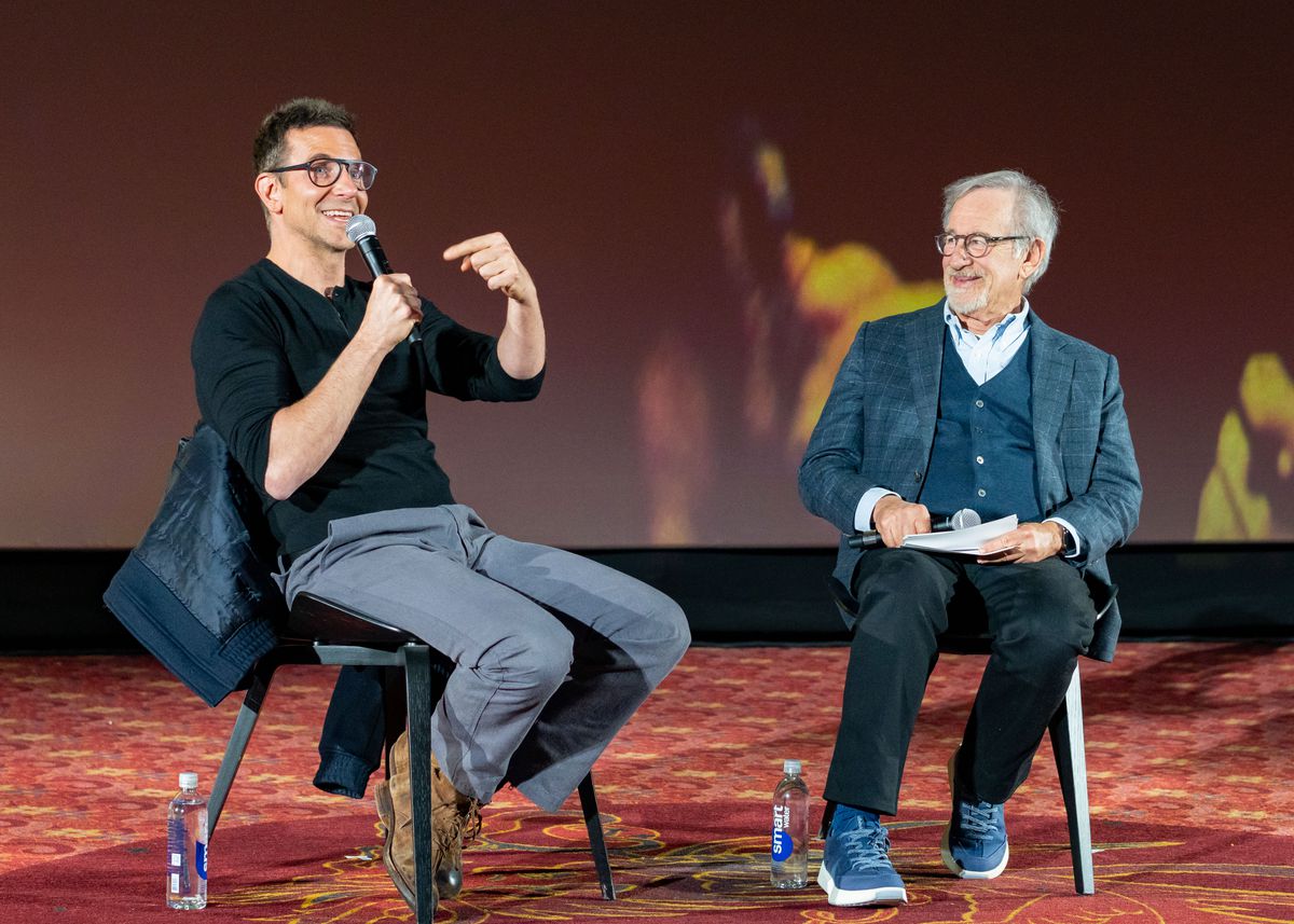 Steven Spielberg interviews Bradley Cooper onstage. Cooper is holding a microphone and pointing. Both are seated in chairs. At the TCL Chinese Theatre on December 9, 2023, in Los Angeles, California.