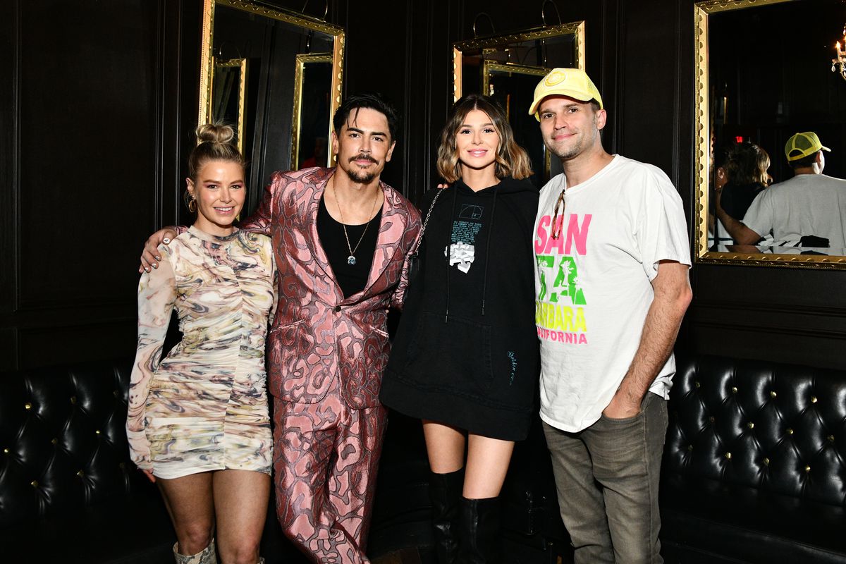 Four people, two men and two women, stand arm and arm, smiling and posing for a photograph. Tom Sandoval wears a satin rose-pink suit and black shirt; Rachel Leviss wears a black mini dress with knee high boots; Ariana Madix wears a form-fitting beige mini dress; Tom Schwartz wears a baseball cap and t-shirt.