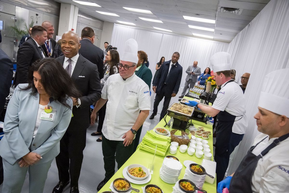 Chefs wearing tall white chef hats and black aprons serve food from buffet tables covered in lime green tablecloths. Mayor Eric Adams walks past, smiling, beside a chef wearing black-rimmed eyeglasses. 