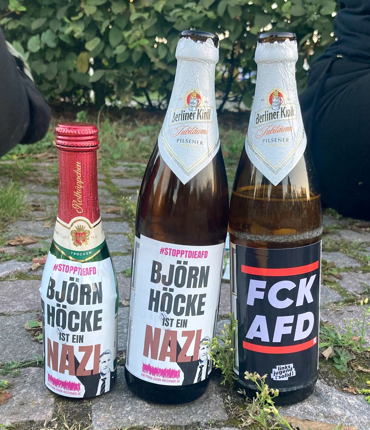 Three beer bottles, against a backdrop of green plants, have labels equating the AfD party with Nazism.