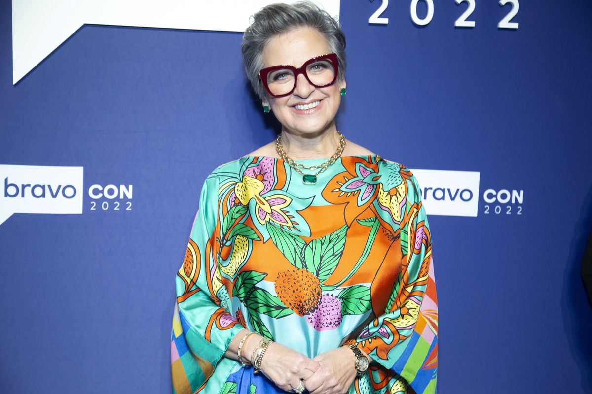 Caroline Manzo poses for a photo in-front of a blue backdrop bearing the words BravoCon 2022 at the Manhattan Center in New York City on October 14, 2022. She has short gray hair, and wears large emerald and gold jewelry, statement eyeglasses with maroon frames, and a silk blouse with a bold orange, pink, turquoise, and yellow floral print.