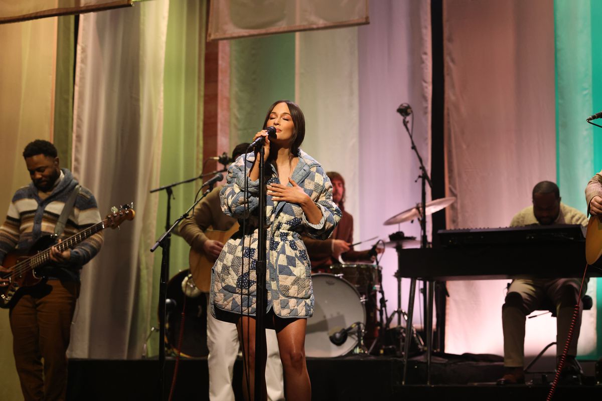 Kacey Musgraves, in a white and blue quilt-patterned dress, sings on stage with a group of musicians backing her.