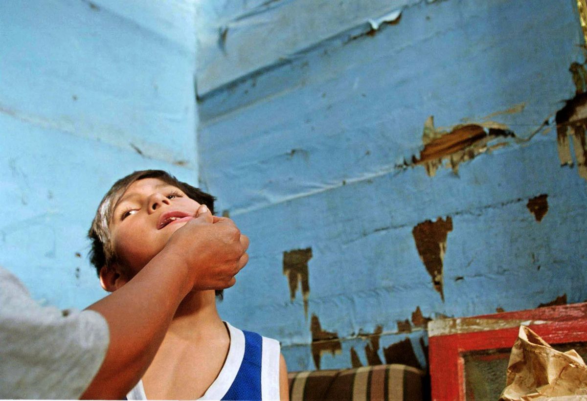 A child in front of a wall with peeling paint leans their head back as a hand touches their face.
