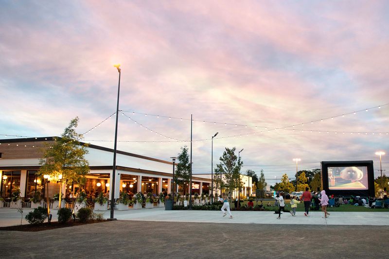 Children are running playfully toward a restaurant with outdoor seating and box planters surrounding. There are strung lights and a paved walkway in front of an outdoor movie screen.