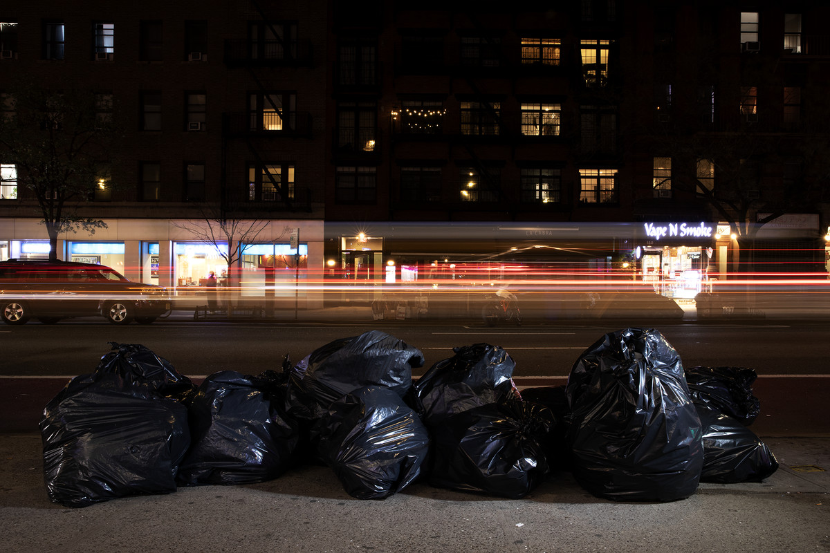 Black trash bags are piled on a city sidewalk, with traffic a blur of lights passing by.