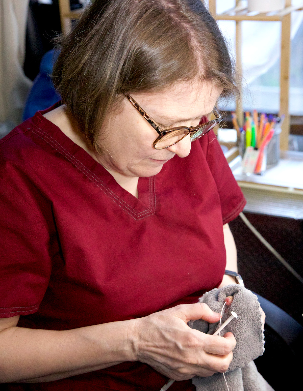 A woman with short brown hair and a red shirt holds a tiny baby opossum in a soft cloth while hand-feeding it through a tube.