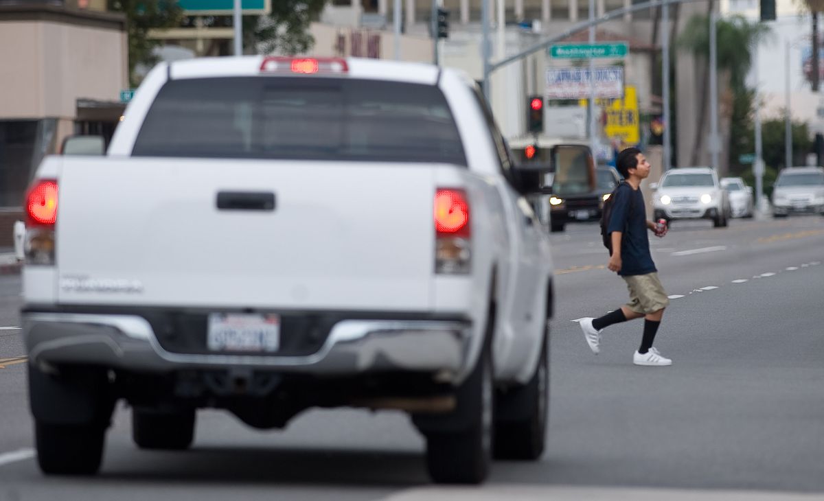 A pedestrian crosses a city street in front of a large white pickup truck in the foreground.