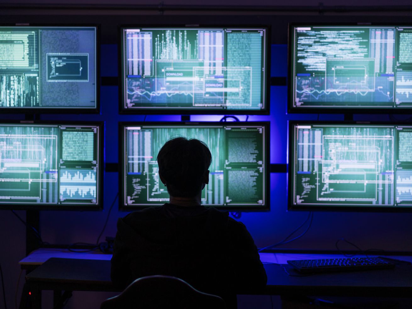 A silhouetted person sitting in front of six computer screens full of data.