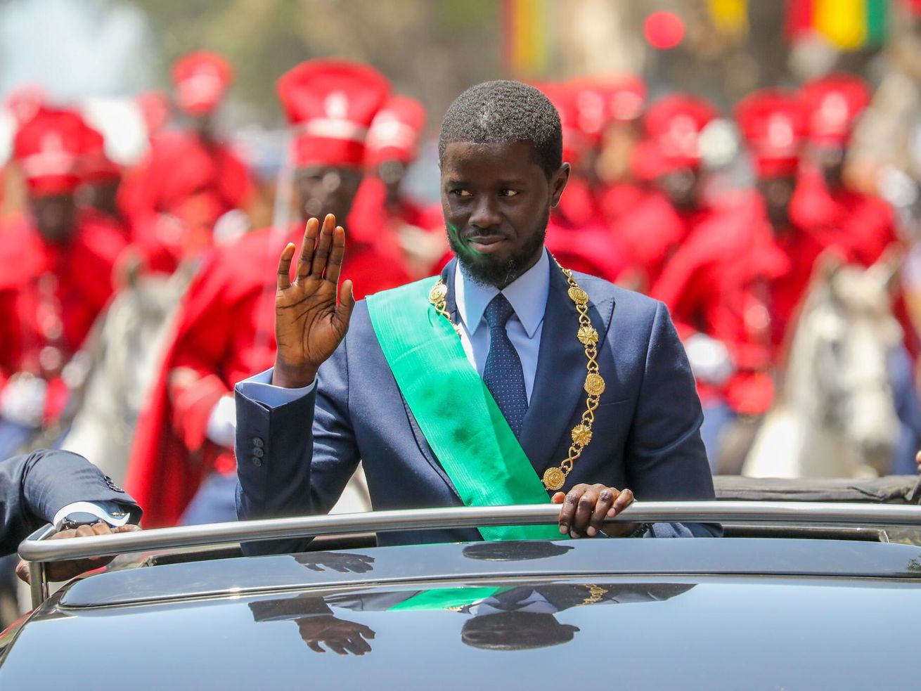 Diomaye, wearing a green sash and suit, stands in an open-roofed vehicle and waves. Behind him, a group of people wearing red ceremonial robes and hats ride white horses.