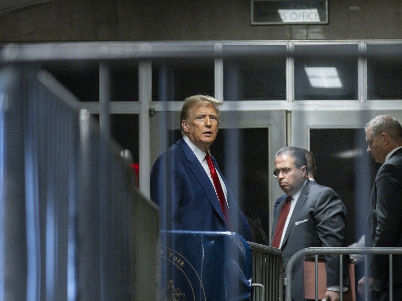 Donald Trump, in a navy suit and red tie, is seen from between barricade bars outside a New York court building, speaking to onlookers.