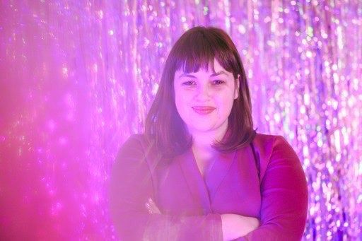 A smiling woman with brown hair stands with her arms crossed in front of a sparkly pink backdrop. The photo is infused with pink light.