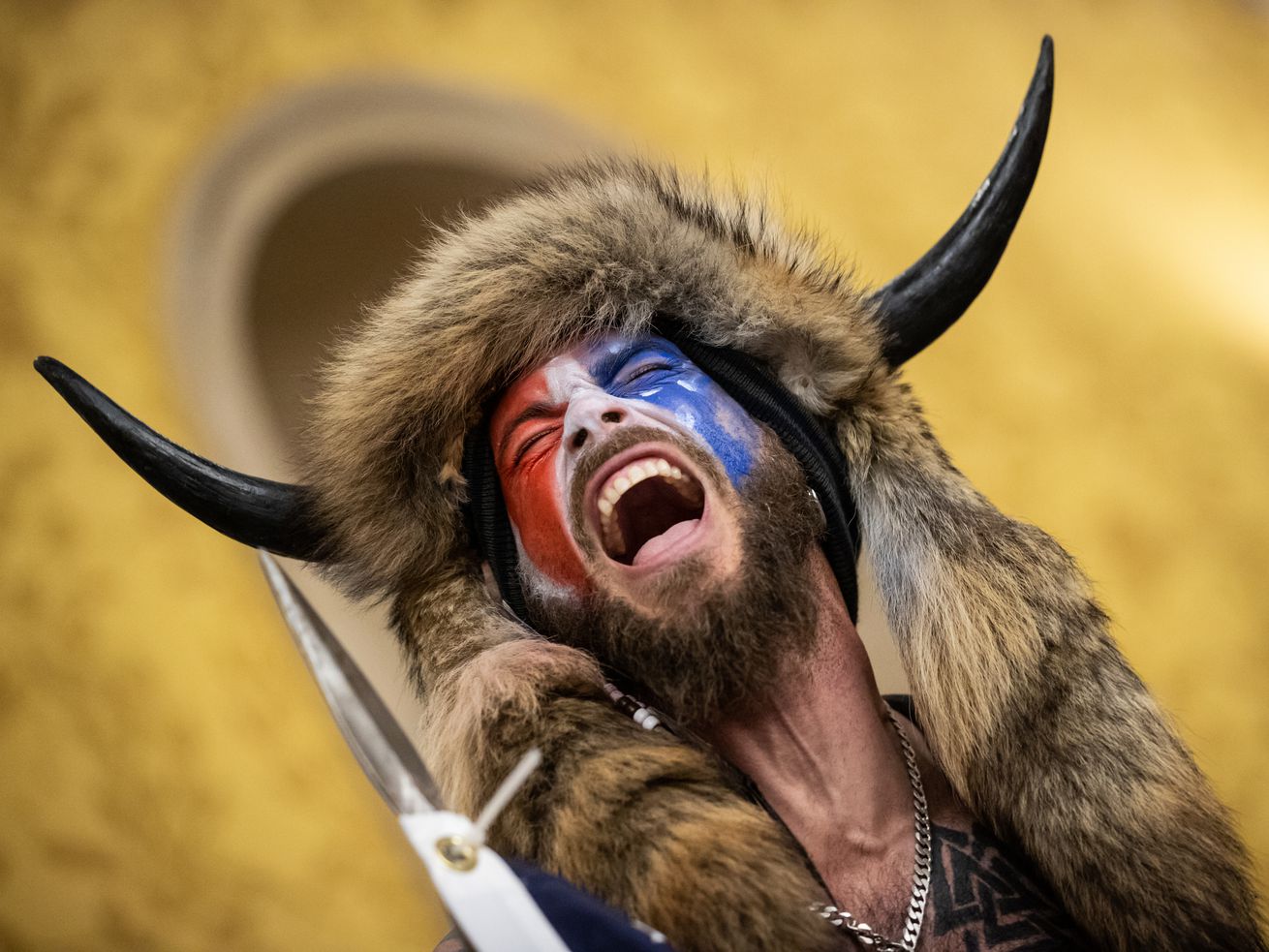 A bearded man in red, white, and blue face paint and wearing a furred and horned hat has his mouth open in a scream.