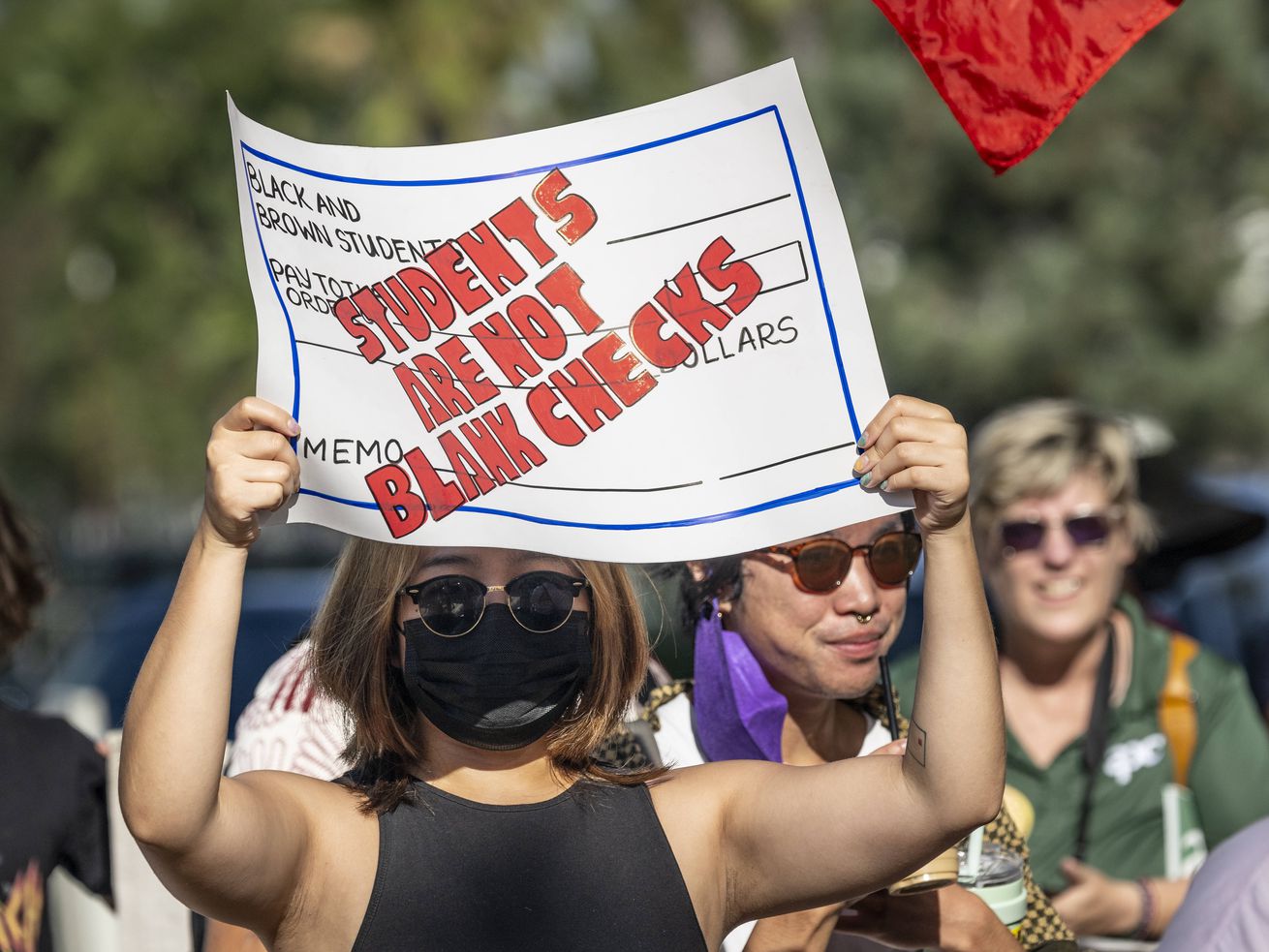 A woman wearing a face-mask and sunglasses holds up a protest sign designed to look like a check that reads “Students are not blank checks” in red letters.