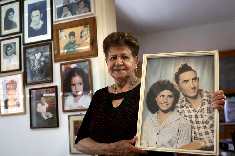 An elderly woman next to a wall full of family photos holds up a portrait of her husband and herself when they were young.
