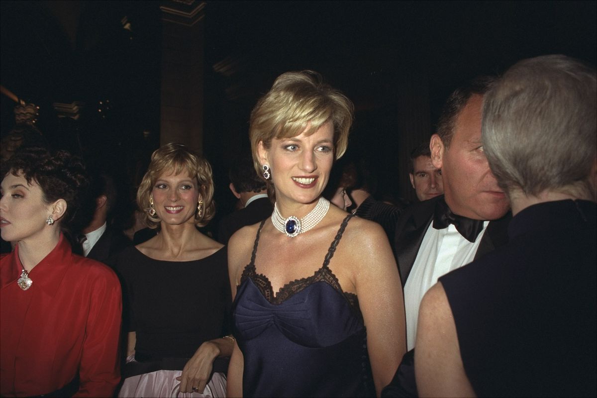 Princess Diana, wearing a dark blue slip dress with black lace trim and pearl-and-sapphire jewelry, smiles at someone in a crowd surrounding her.