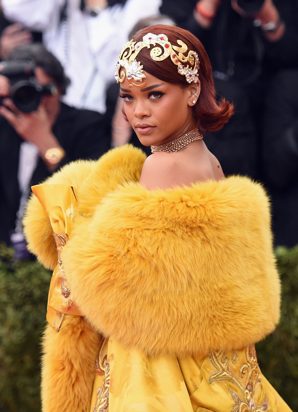 Rihanna, with an elaborate gold headdress and bright yellow fur-trimmed brocade gown, appears at the Met Gala before a crowd of onlookers.