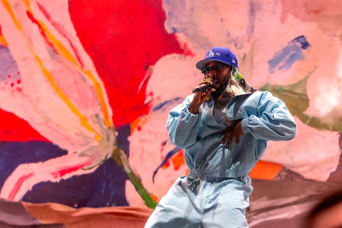 Kendrick Lamar in track suit and Dodgers hat, performing onstage in front of a painted mural of large colorful flowers.