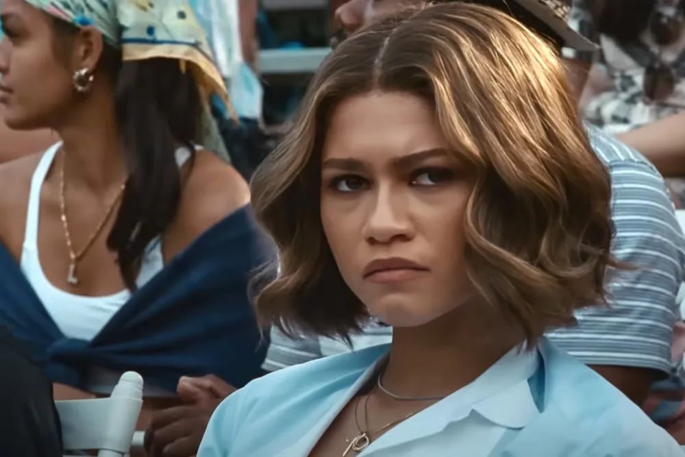 Zendaya as Tashi Duncan in Challengers, wearing a frown and sitting amid a crowd of people.