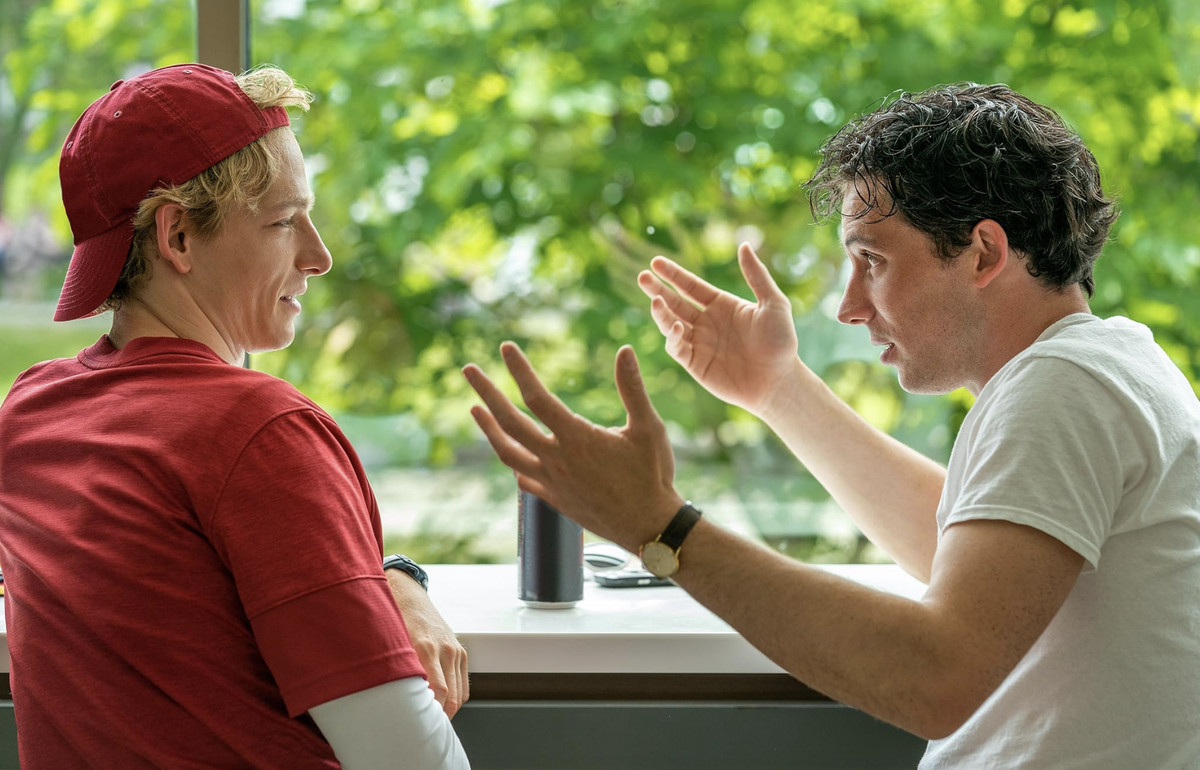 Two young men sit at a table, turned toward each other, while the one on the right gestures with both hands. 