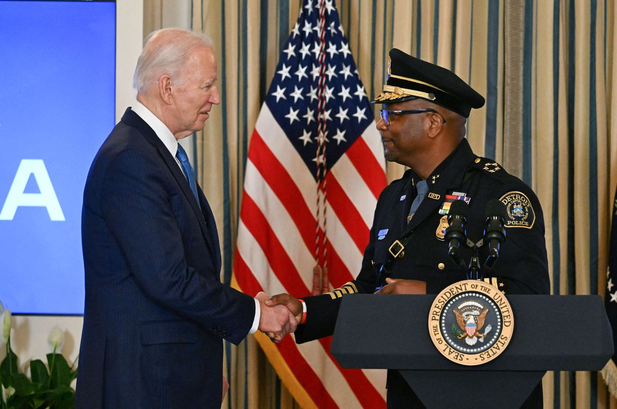 Biden, wearing a suit and tie, and White, wearing a decorated police uniform and hat, shake hands beside a podium and an American flag.