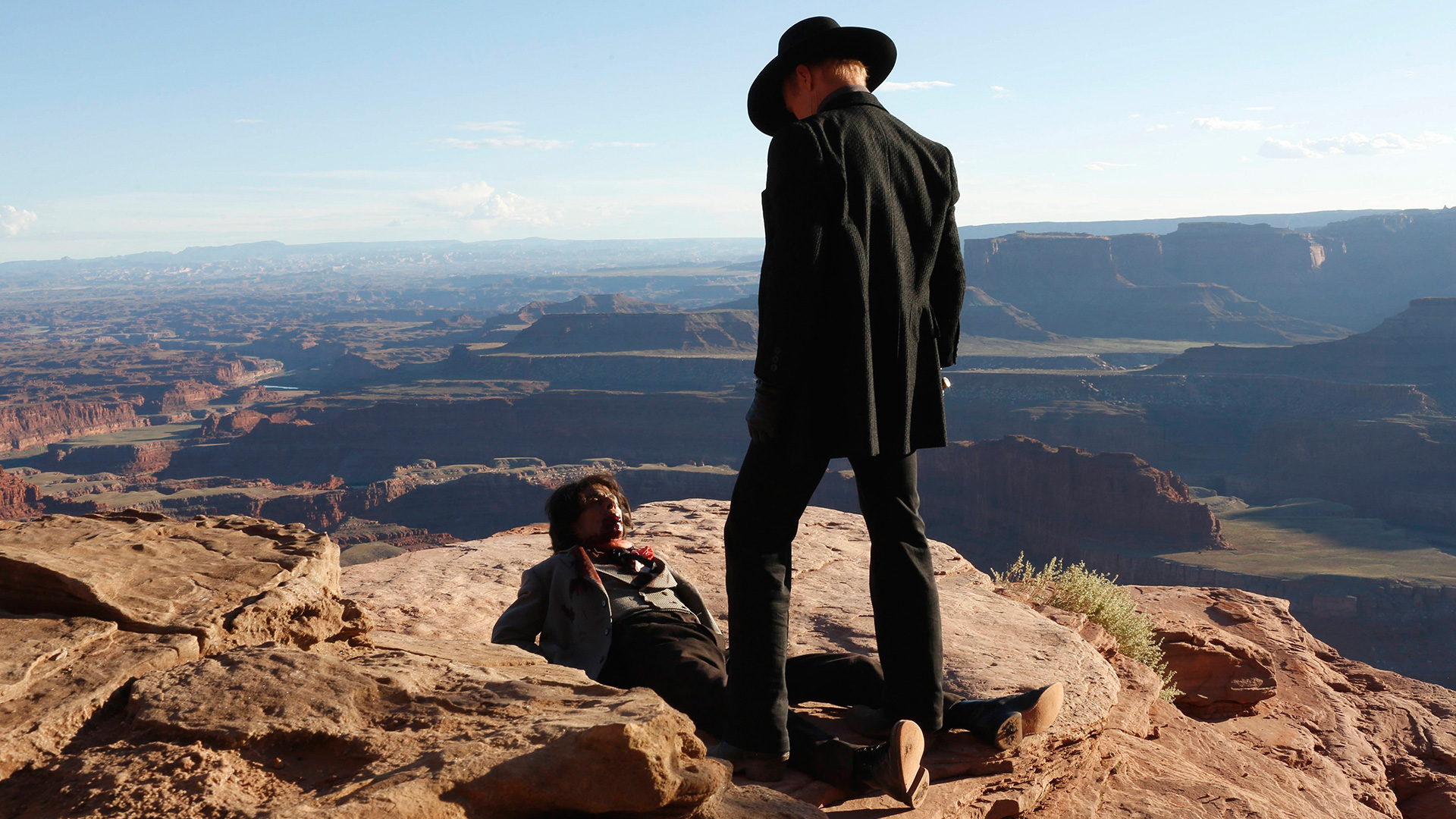 A man in a black suit and cowboy hat stands above another man lying on the ground. They are surrounded by a rocky, desert canyon landscape.