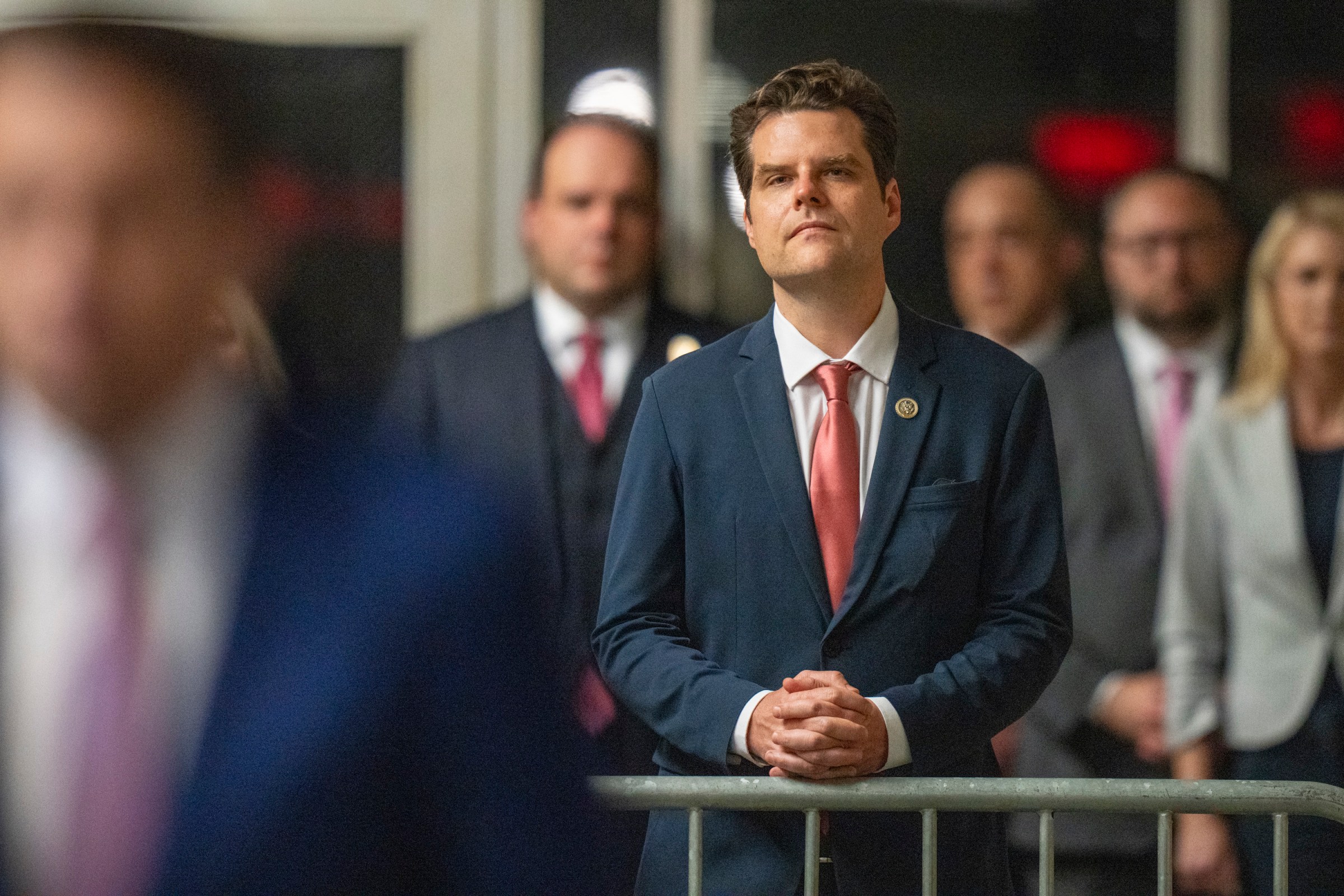 Mat Gaetz standing at a metal barricade in a dark suit and red tie.