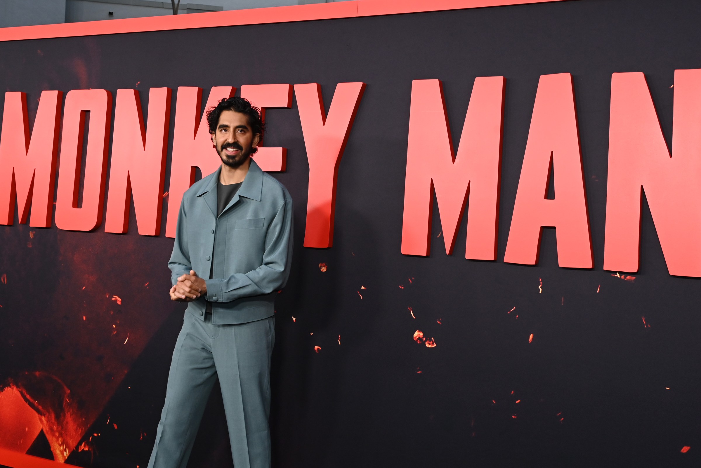 Dev Patel stands on a red carpet in front of the Monkey Man logo.