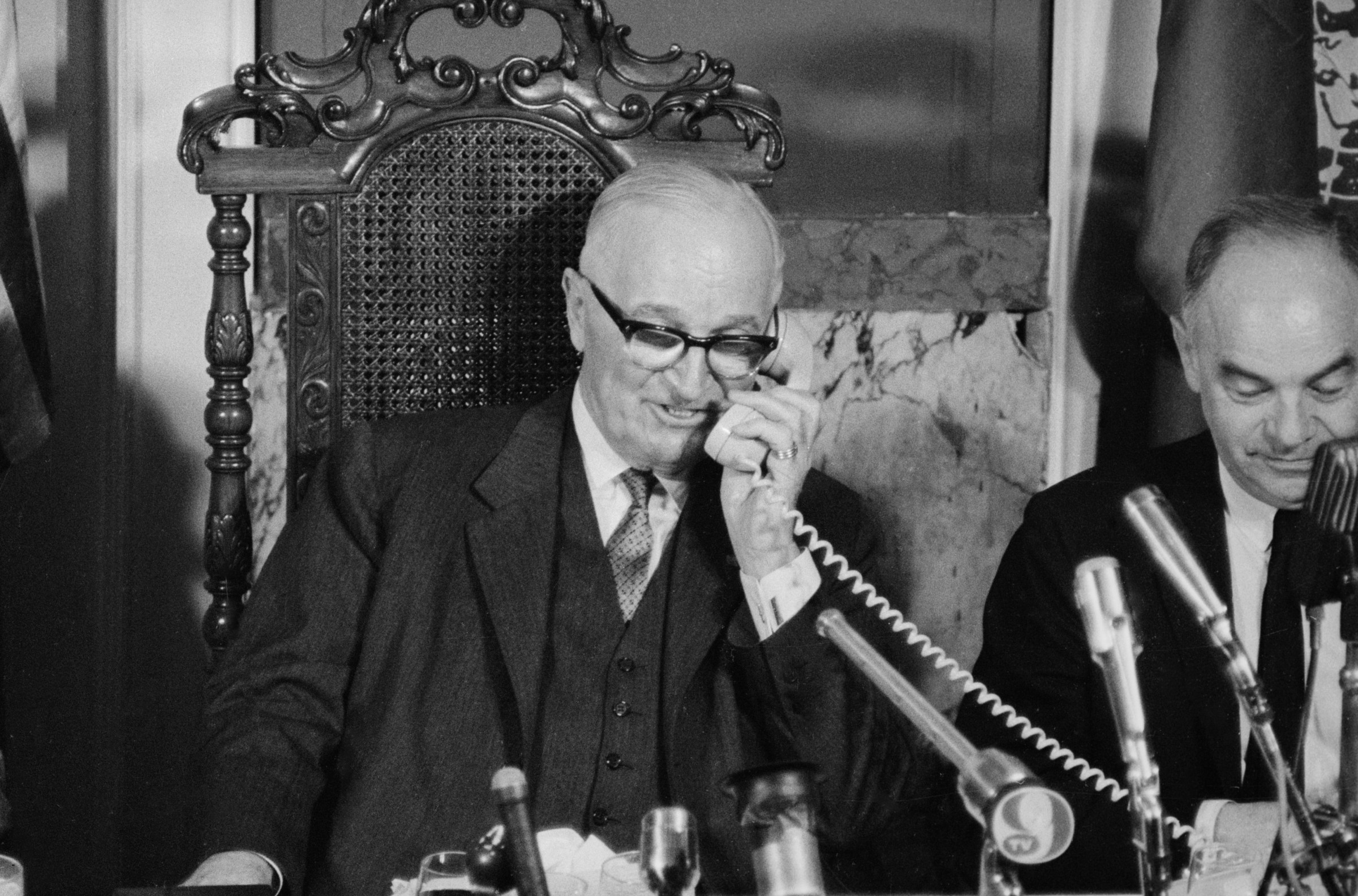 Harry Truman sitting at a desk holding a corded phone receiver to his ear.