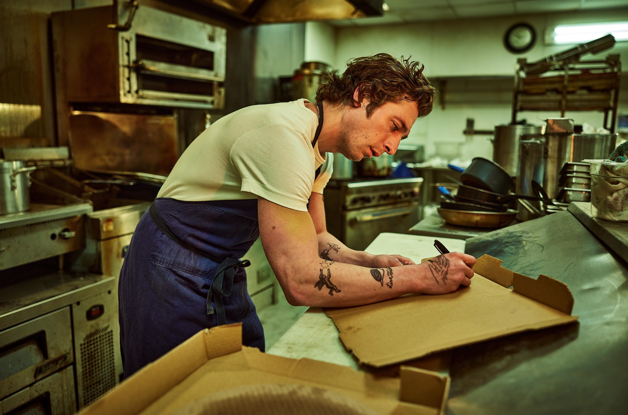 A tattooed man in a white T-shirt and blue apron leans over a kitchen counter, writing on a piece of cardboard.