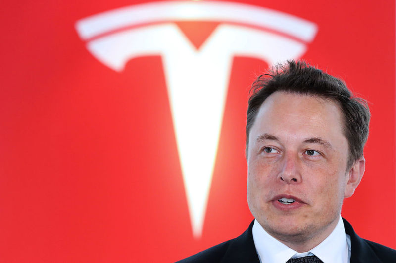 Elon Musk speaks in front of a giant Tesla logo.
