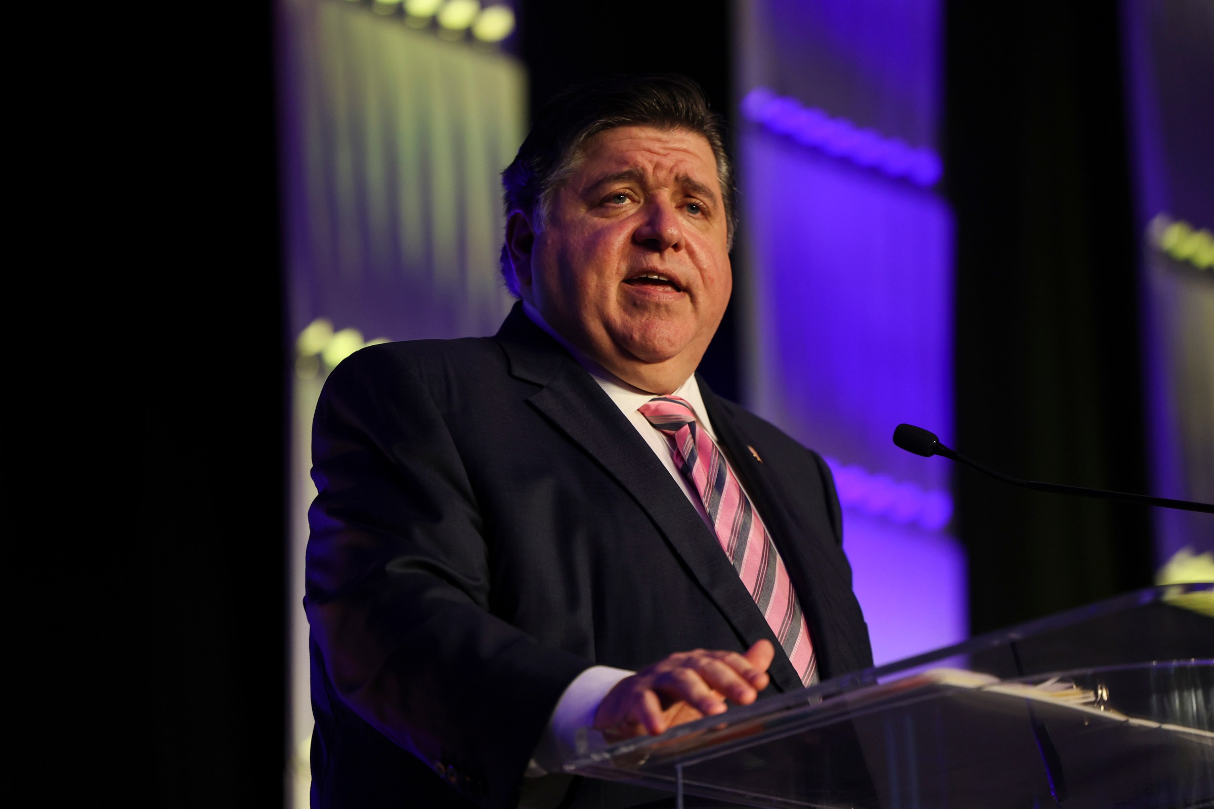 Gov. J.B. Pritzer, in a dark suit and pink striped tie, speaks into a microphone at a podium, with a screen lighting him from the back.