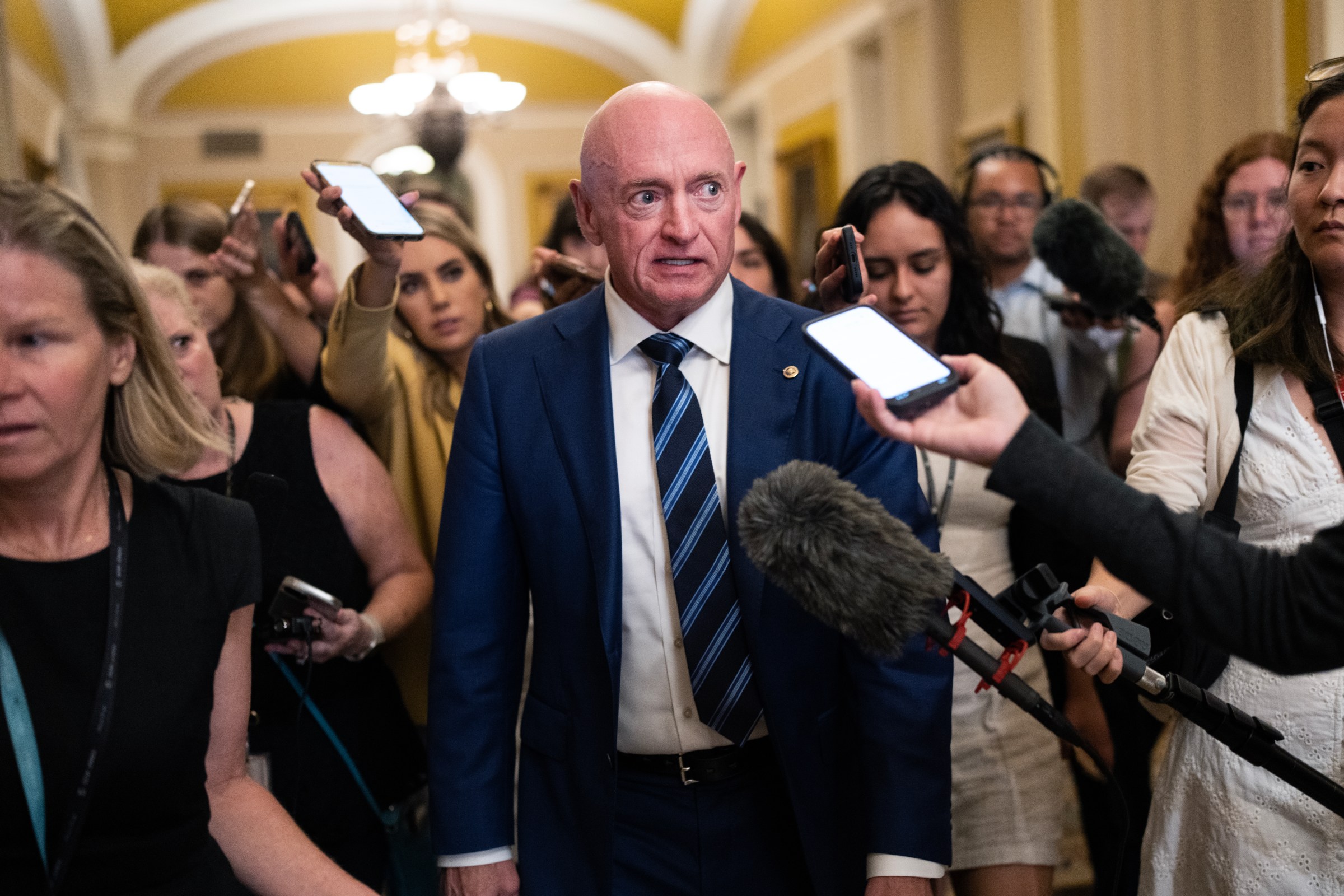 Sen. Mark Kelly, in a navy suit and blue-striped tie, wears a serious expression while speaking to a gaggle of reporters with microphones and smartphones. 