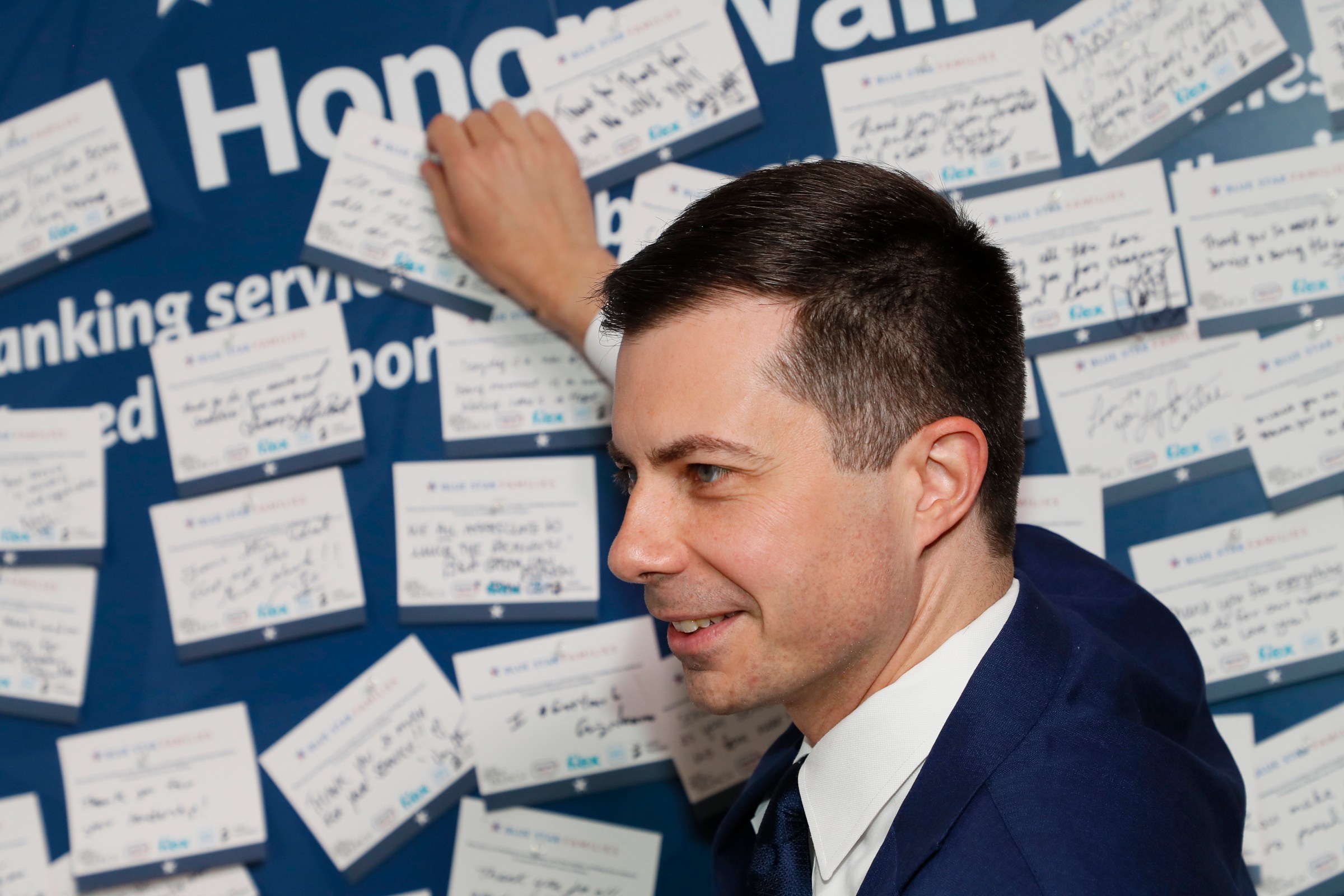 Pete Buttigieg, in a navy suit and white shirt, is shown from the side, while pinning an index card onto a wall full of similar cards with handwritten notes. 