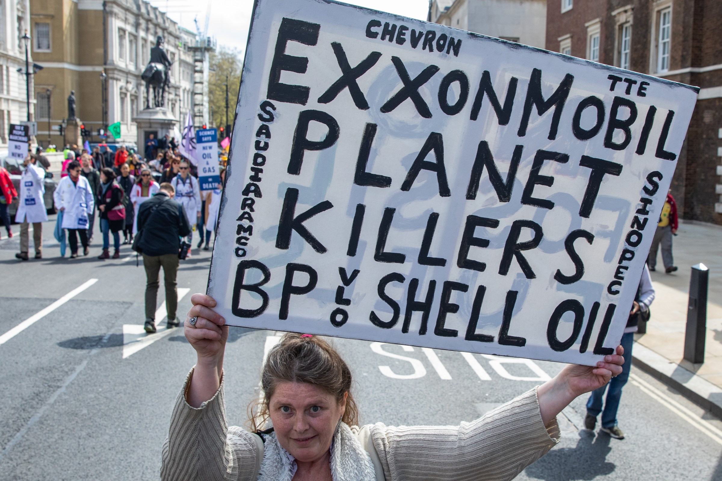 A woman walking in a London city street holds a large sign reading “ExxonMobil Planet Killers, BP, Shell Oil