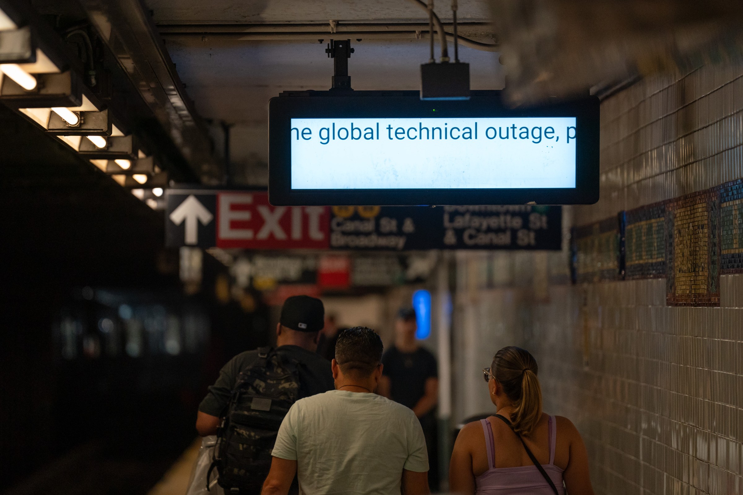 A monitor screen in a subway station reads “the global technical outage,” as people walk underneath it in the direction of signs pointing to the exits.