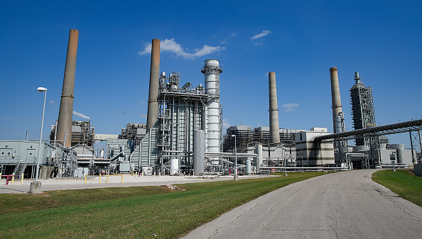 Under a blue sky and at the end of a long asphalt road stand many power plant towers and walkways in a large energy complex.