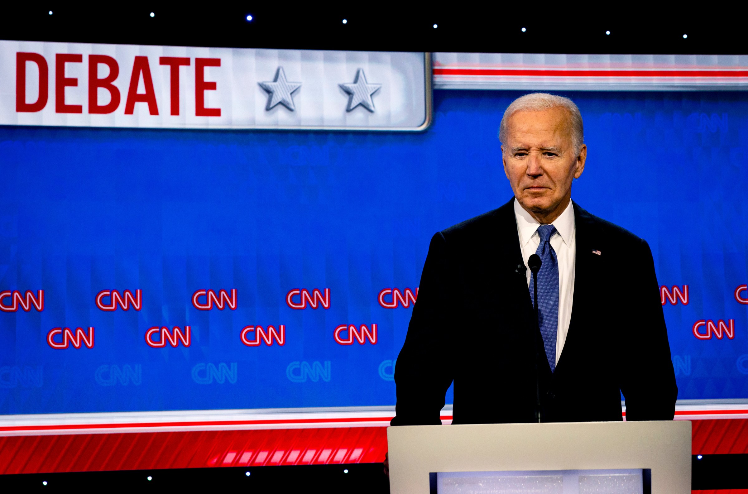 President Biden, in a dark suit against a bright blue background with “CNN” repeated across it, stands at a podium speaking into a microphone.