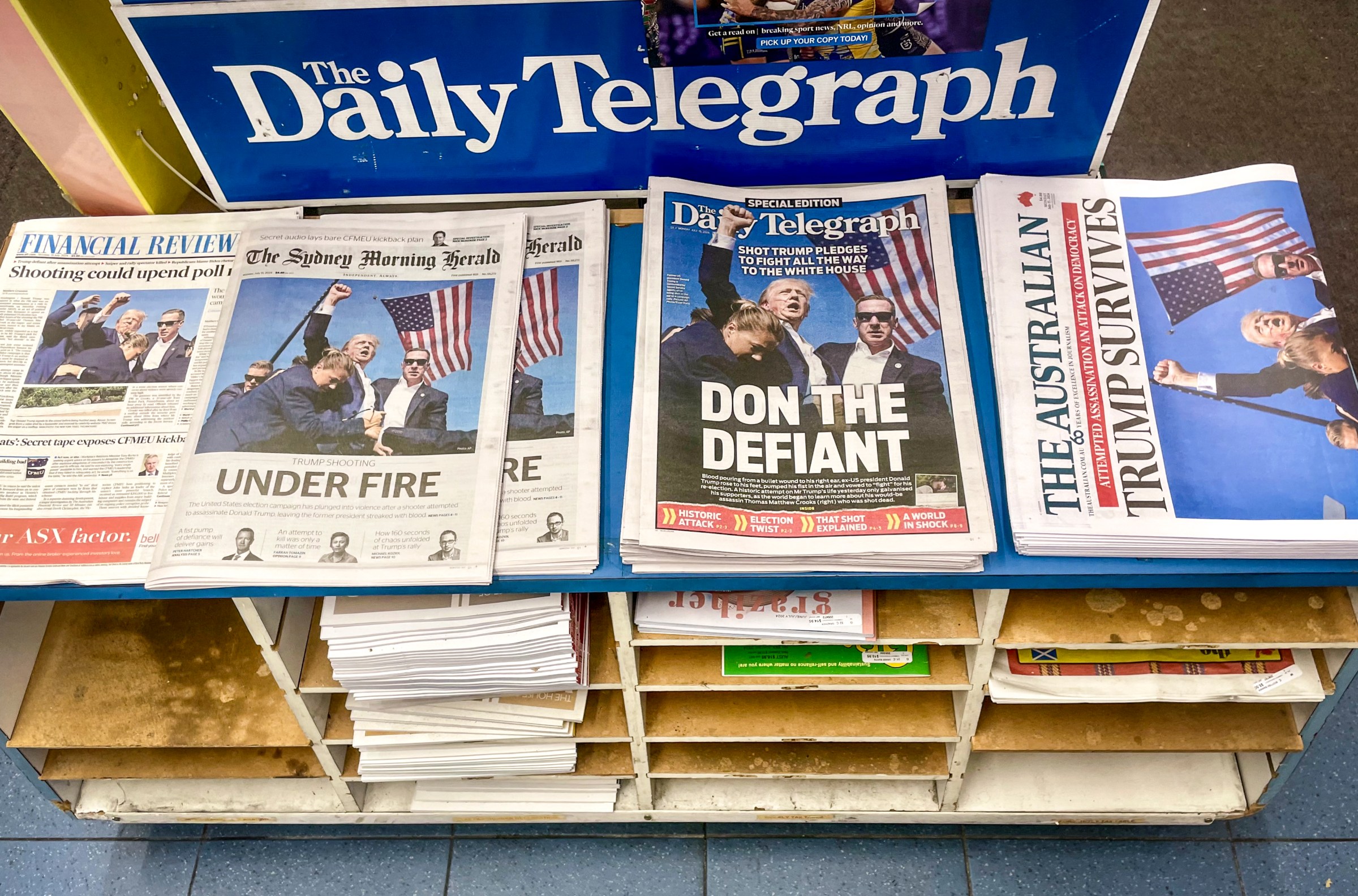 Several Australian newspapers sit in stacks on a wooden shelf with "The Daily Telegraph" on a sign above it. The headlines include "Under Fire," "Don the Defiant" and "Trump Survives." All feature a photo of Trump, fist high in the air, surrounded by Secret Service agents with blood running down the side of his face.