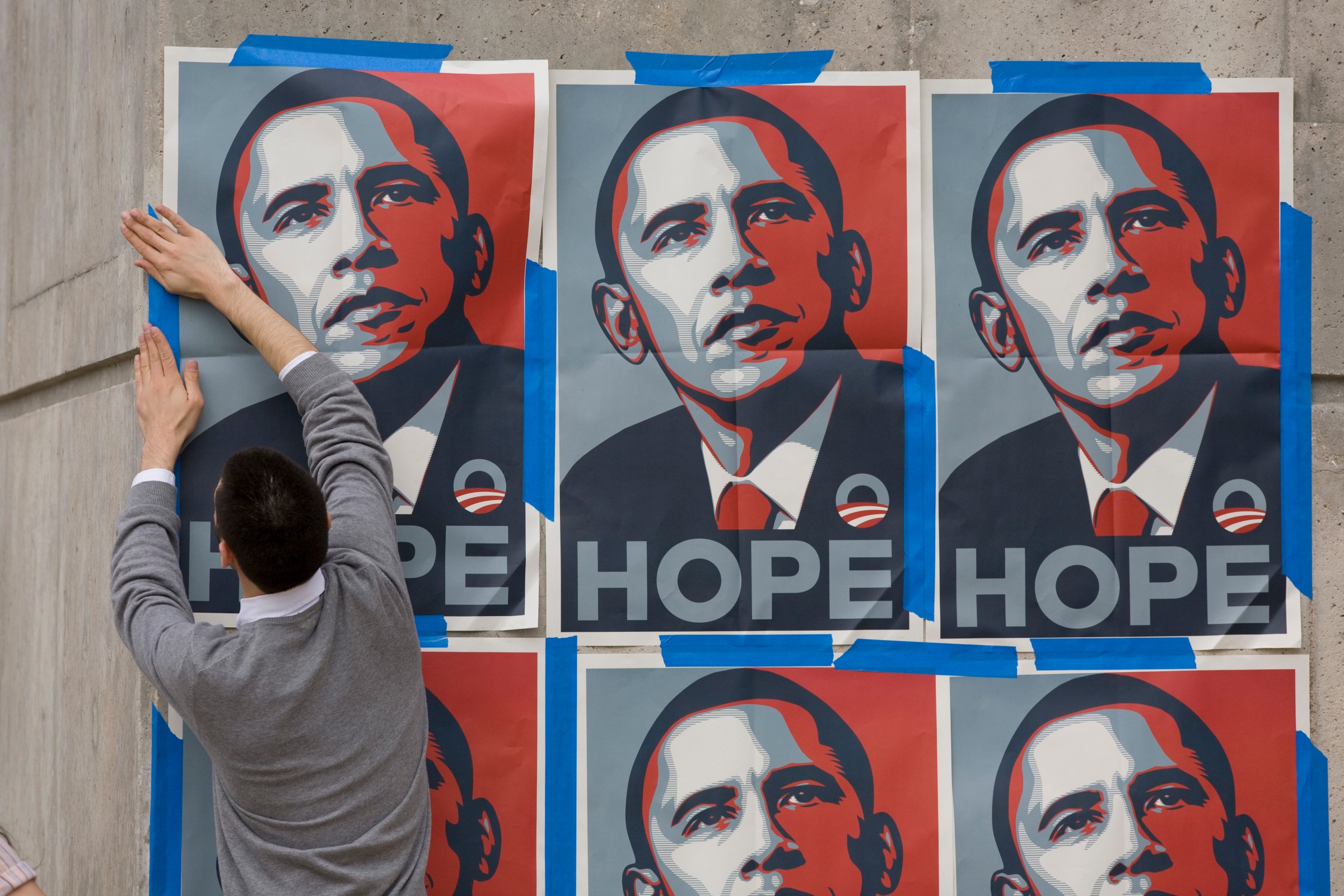 A young man in a gray sweater and white shirt puts up six red, white, and blue posters of Barack Obama’s face with the word “Hope” written below it.