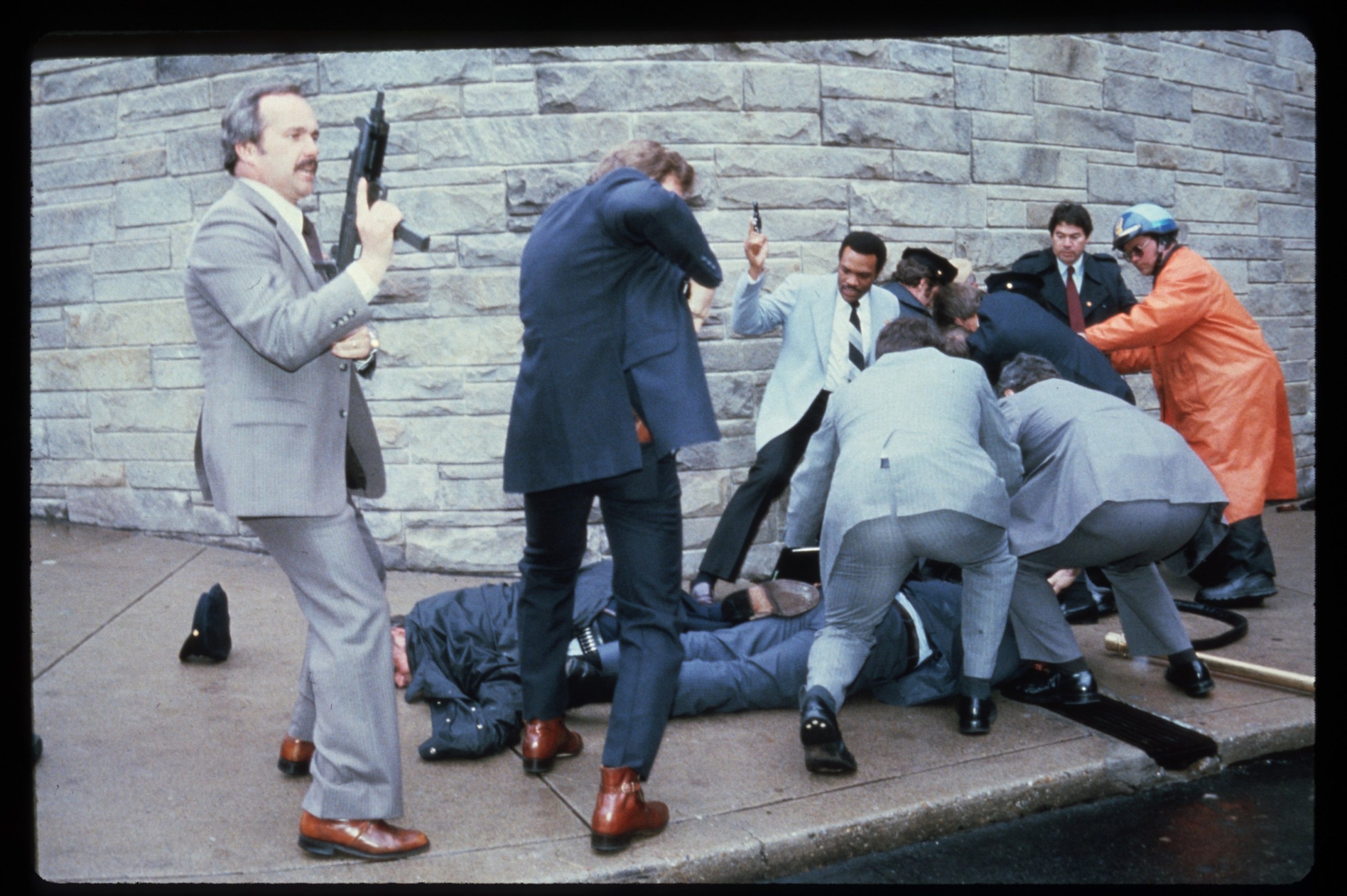 In a photo from 1981, agents in navy and gray suits with guns surround two men who lie on a sidewalk.