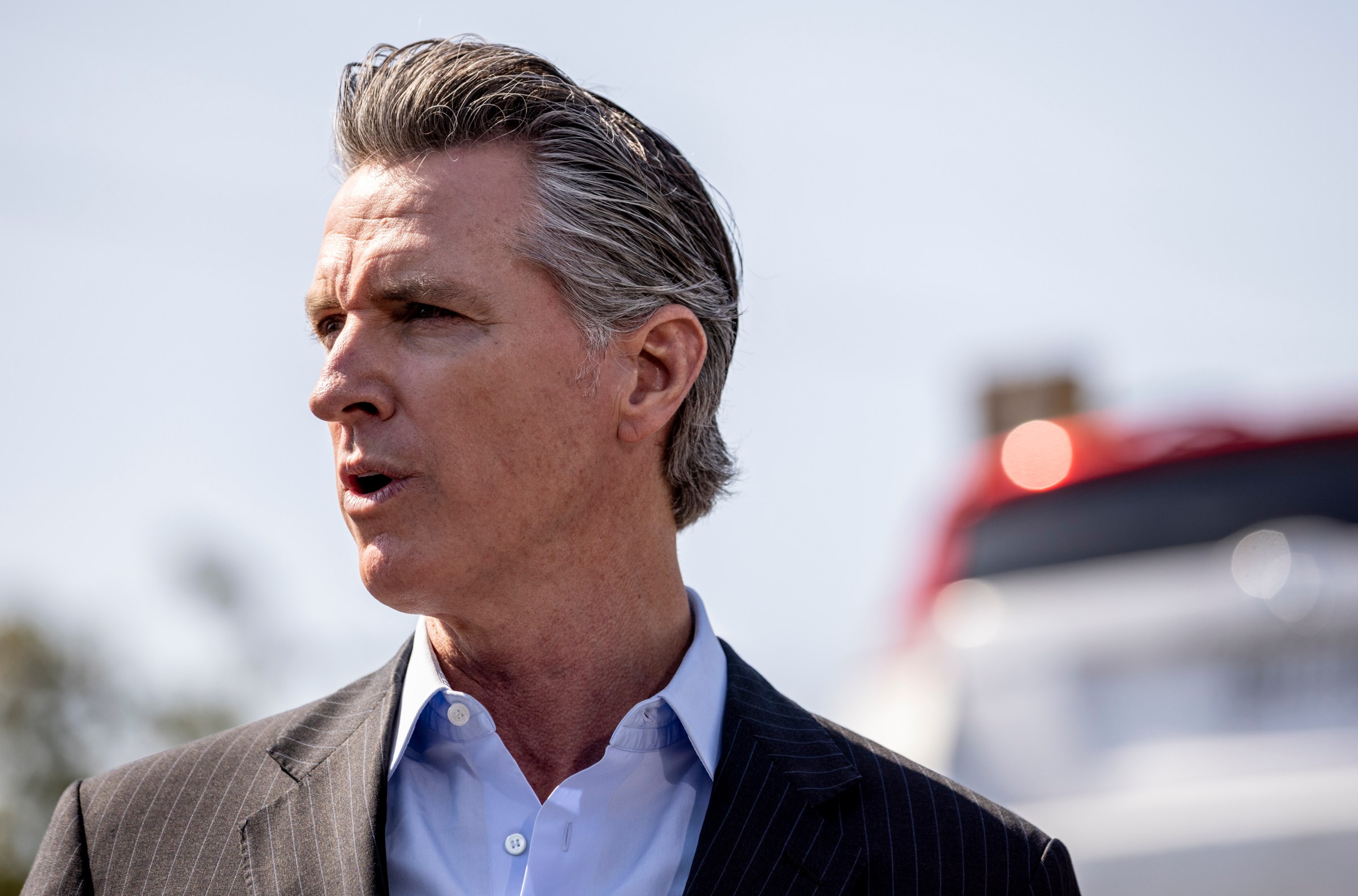 Gov. Gavin Newsom, in a gray striped jacket and blue shirt, squints into the sun as he speaks outdoors.