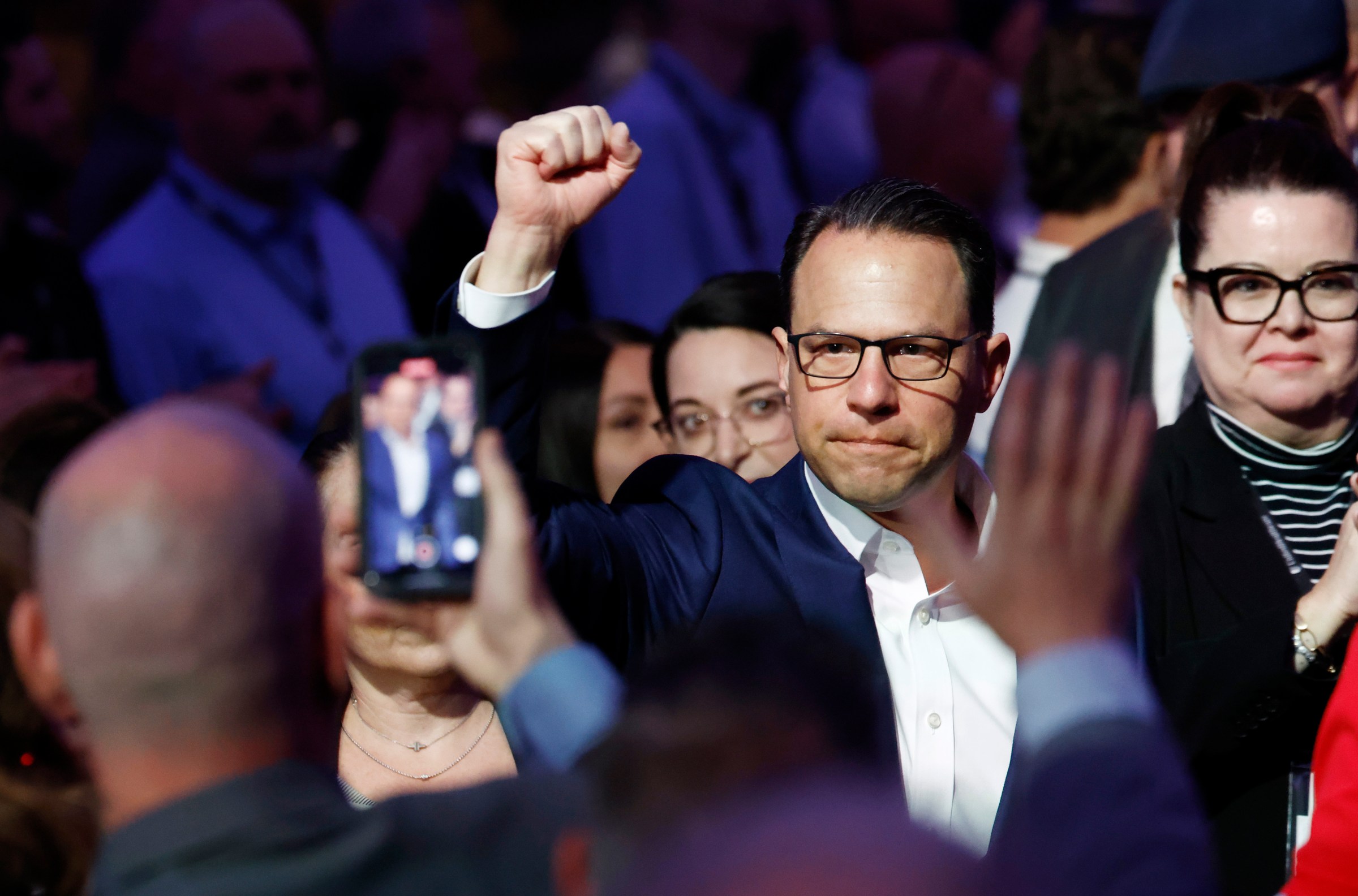 Gov. Josh Shapiro, in glasses and a navy suit jacket, waves at reporters and a crowd.