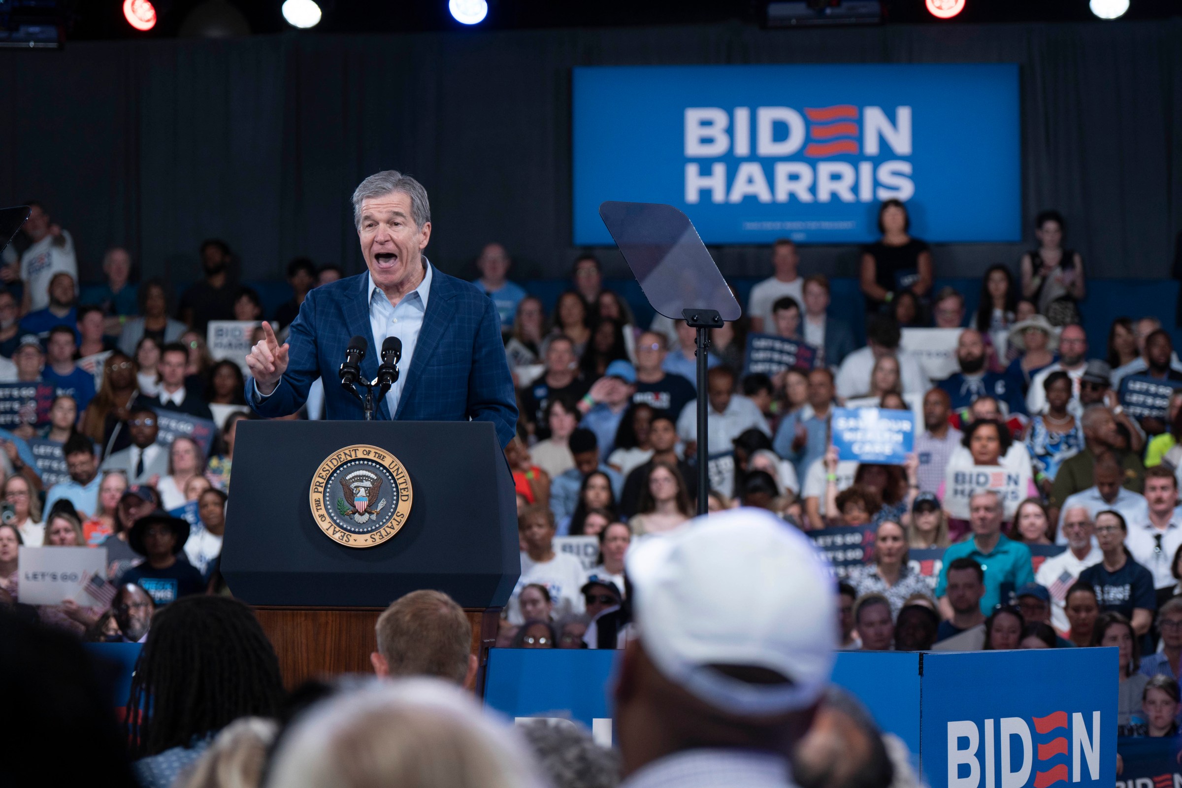 Gov. Roy Cooper, in a navy suit, speaks from a podium to a large crowd holding Biden-Harris campaign signs.