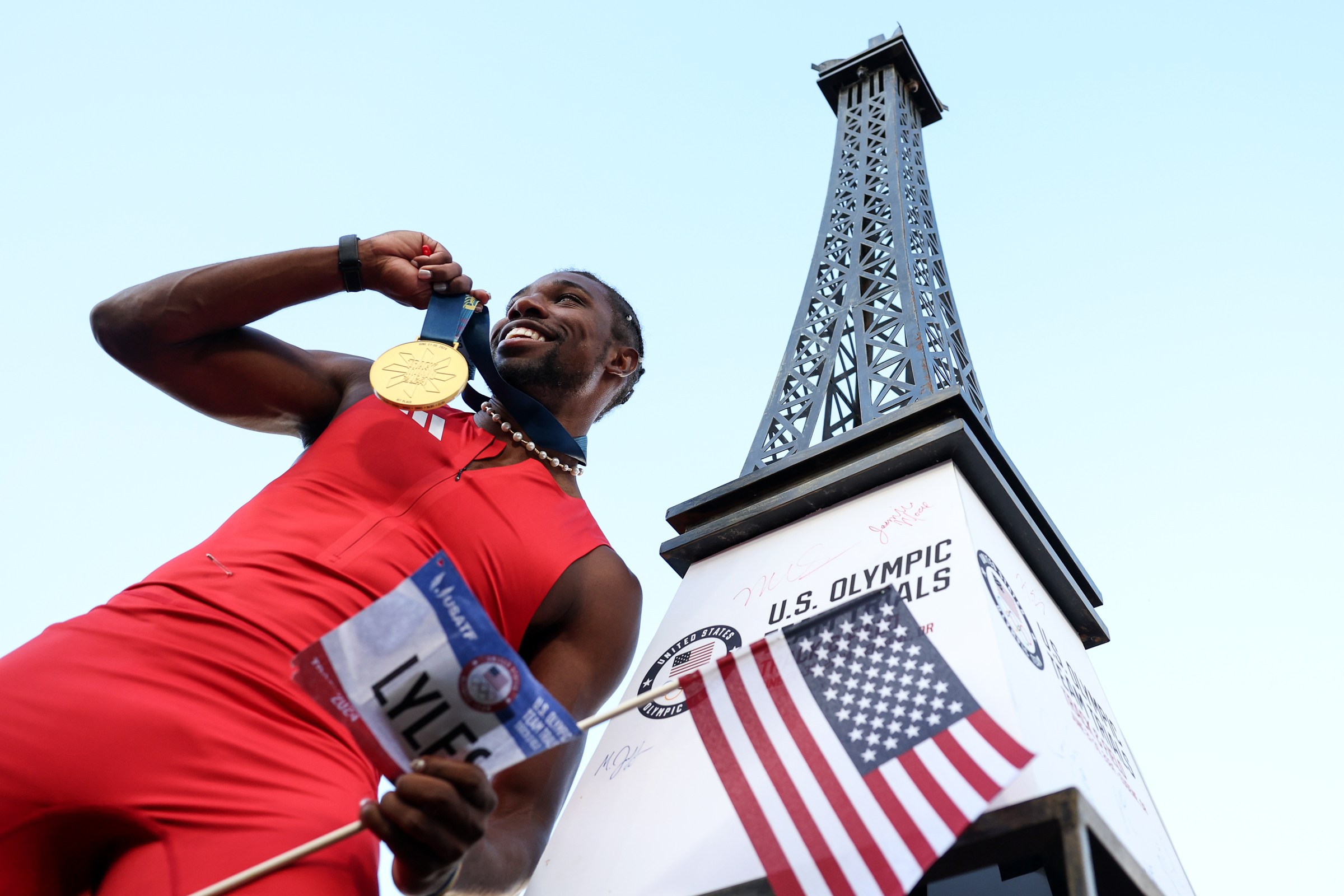 Noah Lyles, pictured from below, wears a red track suit and a gold medal around his neck, stands beside a replica of the Eiffel tower.