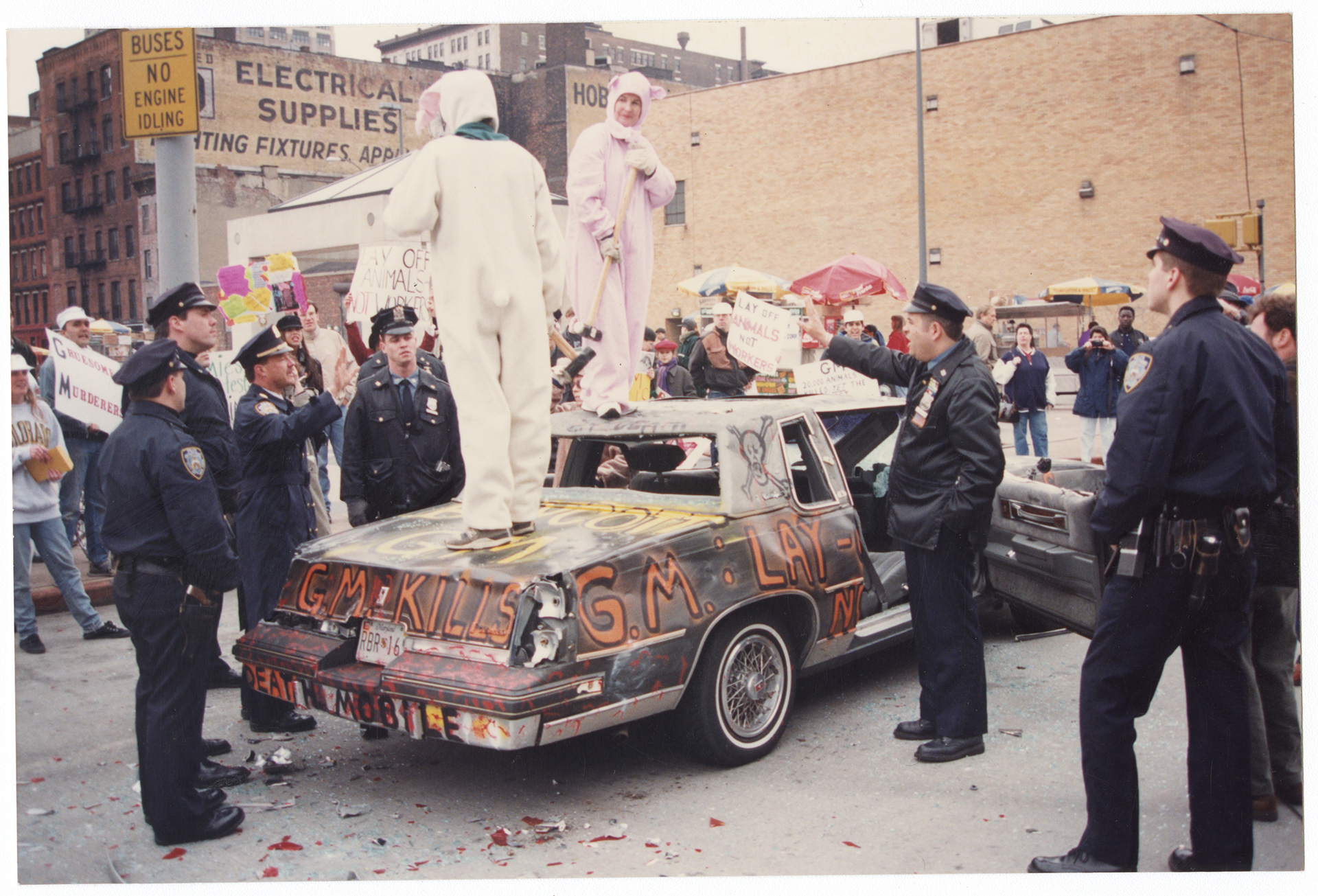 Protesters with sledgehammers dressed in pig costumes stand on top of a GM car with its windows broken, while police engage them and a larger crowd of protesters stands around.