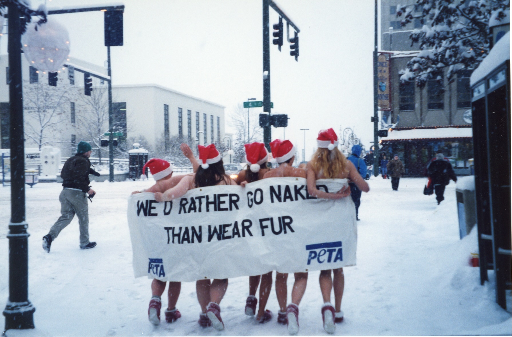 photo of a snowy street with a view of four activists from behind that appear naked, each wearing Santa hats and holding a large banner behind them that reads “WE’D RATHER GO NAKED THAN WEAR FUR.” 