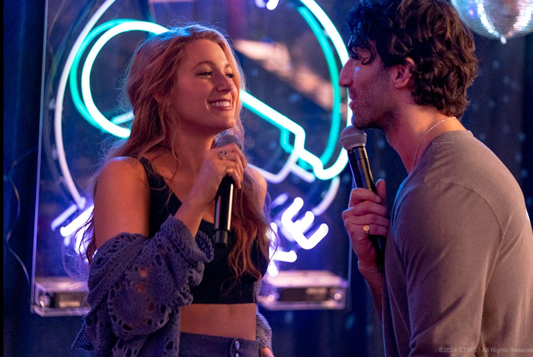 A man and a woman singing karaoke stare at each other with flirty smiles.